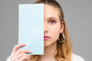 Woman covering half of her face with paper seeking causes of uneven eyes